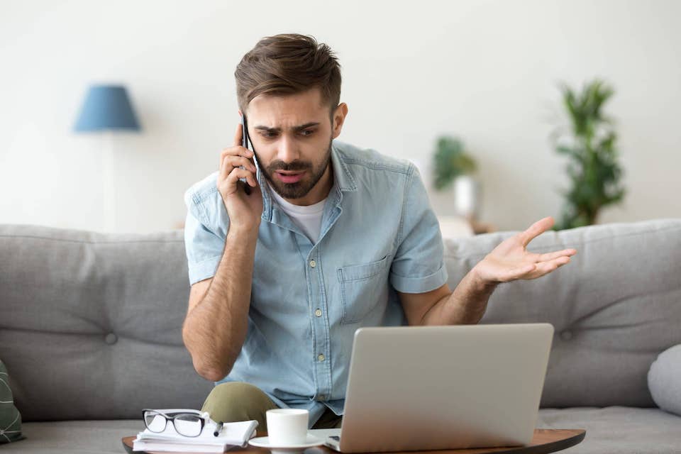 frustarted man on phone sitting on the couch with a laptop in front of him; diy roofing 