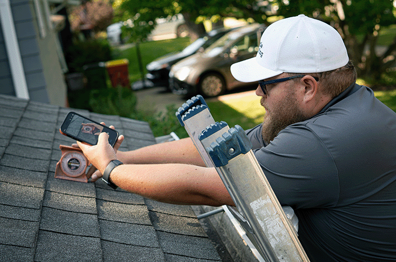 A roofing consultant measures roof pitch with a gauge while standing on a ladder against an old and worn shingled roof. The consultant is taking a photo for documentation.