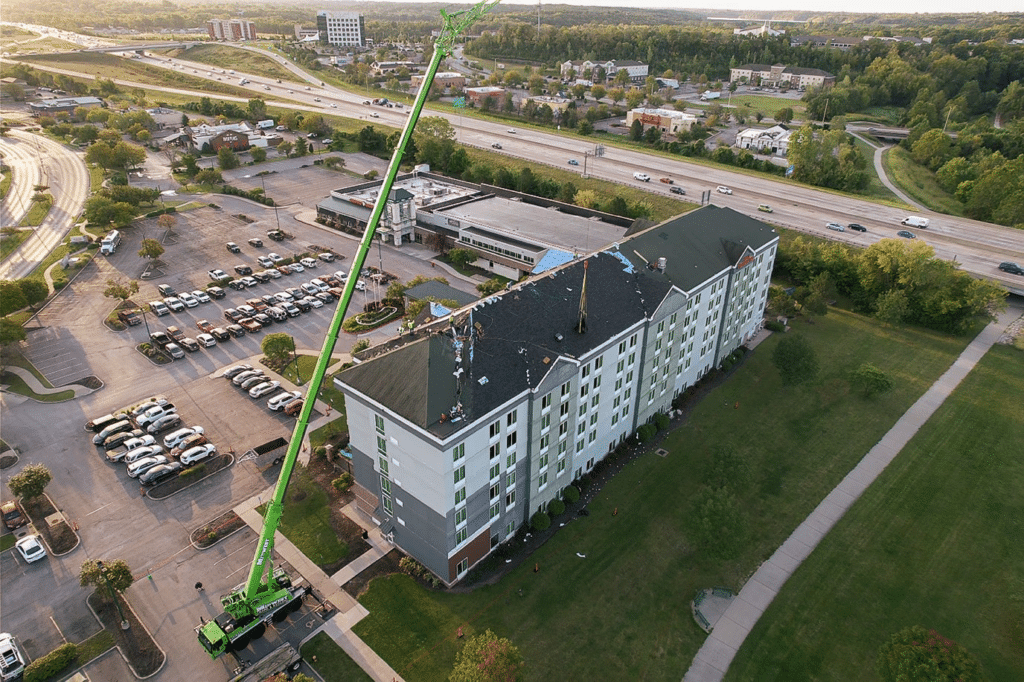 Aerial view of Hilton Garden hotel in Kansas City, Missouri undergoing a commercial roof replacement. A large crane is positioned next to the 7-story hotel, lifting charcoal-colored shingles to match the light grey and navy-grey panels. The hotel is located near a busy highway.