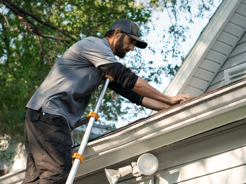 An Apple Roofing employee, conducts a fall roof inspection. He stands on a ladder, reaching out to touch the shingles, ensuring their stability and condition before winter.