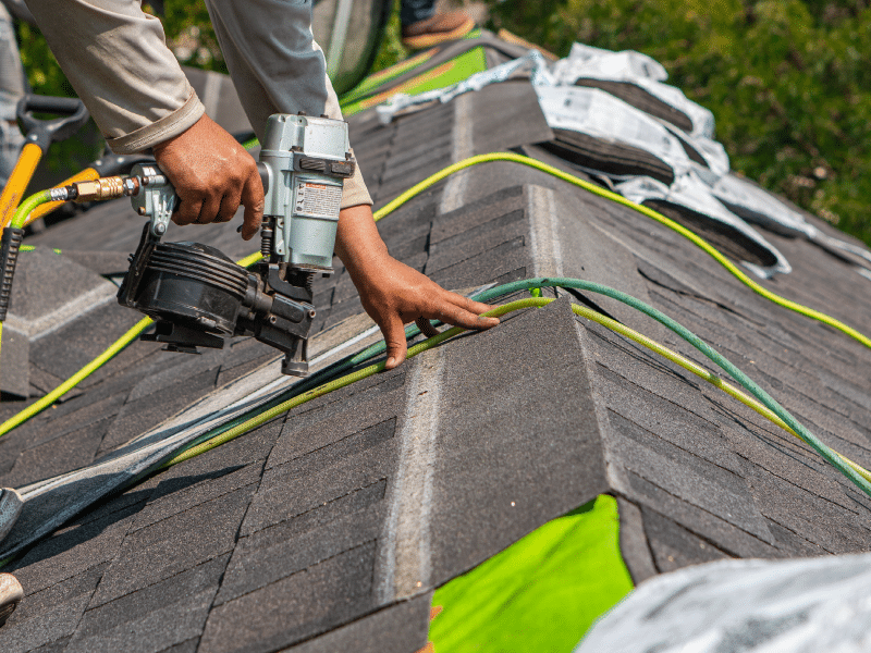 The image shows shingles being installed on a residential roof. In the foreground, a roofer's hands hold a nail gun, ready to secure the shingles to the roof. In the background, open bundles of shingles are stacked next to each other.