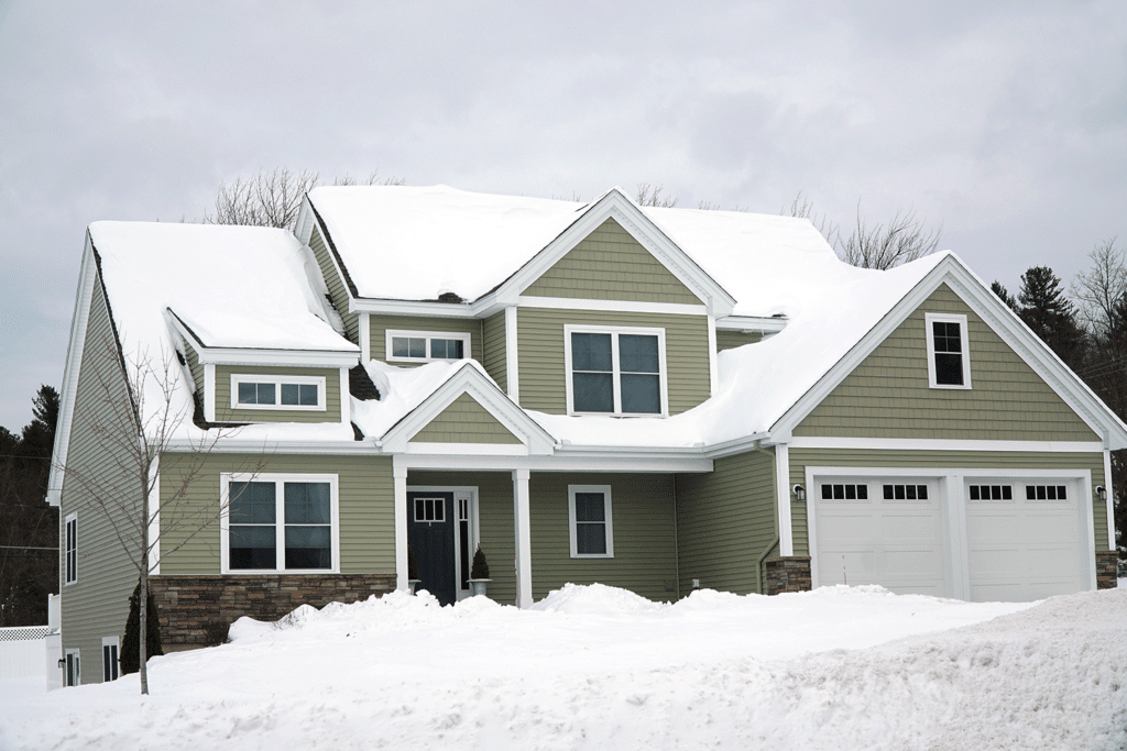 A muted green home stands covered in snow, with mounds of snow blanketing the front lawn and a thick layer covering the roof.
